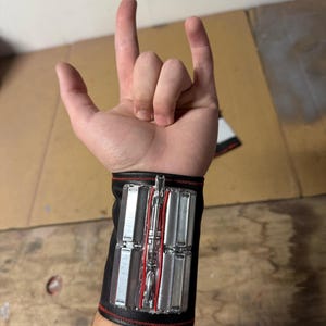May include: Black wrist cuff with silver and red accents. The cuff features rectangular silver panels and a central red design. The cuff is worn on a wrist, with the hand making a rock and roll gesture.