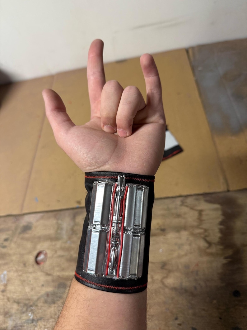 May include: Black wrist cuff with silver and red accents. The cuff features rectangular silver panels and a central red design. The cuff is worn on a wrist, with the hand making a rock and roll gesture.