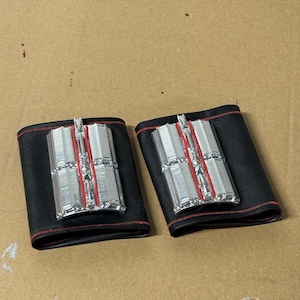 May include: Two black leather wrist cuffs with silver and red accents. Each cuff features a rectangular silver centerpiece with vertical lines and red detailing. The cuffs are stitched with red thread.