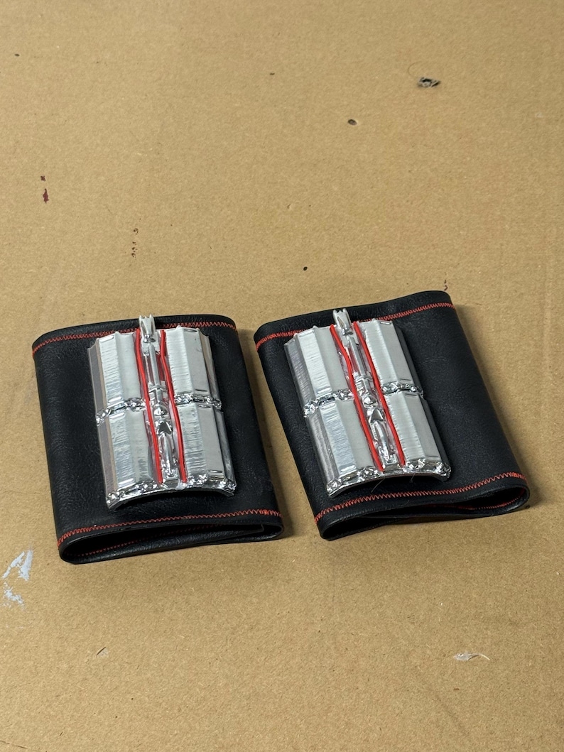 May include: Two black leather wrist cuffs with silver and red accents. Each cuff features a rectangular silver centerpiece with vertical lines and red detailing. The cuffs are stitched with red thread.