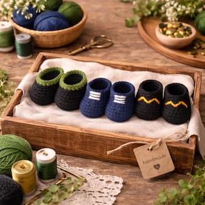 May include: A collection of six handmade baby booties in a wooden tray. The booties are in various colors, including black, navy blue, and green. Some have white or yellow accents. The tray also contains yarn, thread, and a tag that says "handmade".