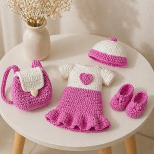 Op de afbeelding: Een gehaakte poppenoutfitset in wit en roze. De set bevat een rugzak, jurk, hoed en schoenen. De jurk heeft een hartapplicatie. De hoed en schoenen hebben roze accenten. De rugzak heeft een witte flap.