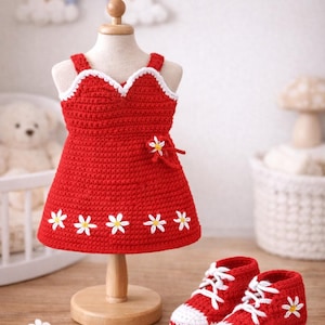 Puede incluir: Vestido rojo de ganchillo hecho a mano con detalles de margaritas blancas y zapatillas rojas a juego con cordones blancos. El vestido se exhibe en un soporte de madera. Un arcoíris y nubes están en el fondo. Un encantador atuendo de bebé hecho a mano.