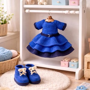 Puede incluir: Un vestido azul de ganchillo hecho a mano con una falda escalonada y cinturón a juego cuelga de una percha de madera. Un par de zapatos azules de ganchillo con cordones blancos se encuentra sobre una pequeña alfombra.