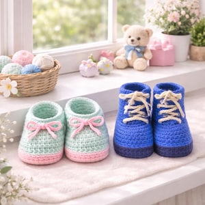 May include: A pair of light blue and pink crochet baby shoes with pink bows, and a pair of blue crochet baby boots with cream-colored laces. The shoes and boots are displayed on a white surface near a window.