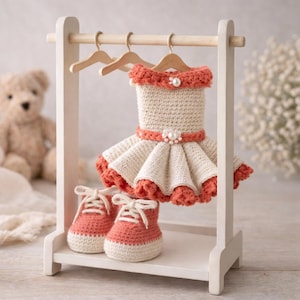 May include: A cream and coral crochet baby dress and shoes set displayed on a small white wooden rack. The dress has a ruffled skirt and pearl embellishments. The shoes are miniature trainers.
