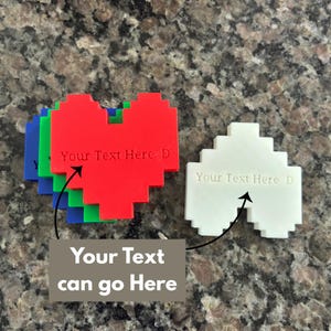 May include: A collection of pixelated heart-shaped ornaments in red, green, blue, and white. Each heart features the engraved text "Your Text Here D" on the front surface. The hearts are arranged in a stack with one white heart placed separately.