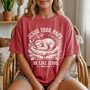 Op de afbeelding: Rood T-shirt met de tekst "JESUS TOOK NAPS" en "BE LIKE JESUS" met een slapende luiaard-afbeelding. Het shirt heeft een relaxte pasvorm en is gemaakt van een zacht materiaal.