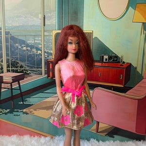 Puede incluir: Muñeca vintage con cabello rojo, vestida con un top rosa sin mangas y una falda dorada con lentejuelas y motivos florales rosas. Un lazo rosa brillante adorna la cintura. La muñeca posa frente a un fondo pintado de estilo retro.