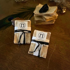 May include: Two stacks of handmade paper, each wrapped with a white label and black twine. The labels feature a logo with a crescent moon and a stylized archway, with the text "MOONHOUSE". A black crystal sits atop a stack of paper.