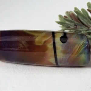 May include: A rectangular glass bead with a hole on each end. The bead features a blend of colors, including brown, purple, yellow, and green, with a black vertical stripe and a black dot. A sprig of greenery is in the background.
