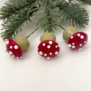 May include: Three red mushroom-shaped glass beads with white polka dots, each with a light green stem. A sprig of green pine needles is in the background. These decorative beads are perfect for jewelry making or craft projects.
