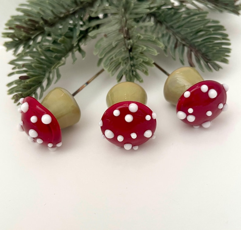 May include: Three red mushroom-shaped glass beads with white polka dots, each with a light green stem. A sprig of green pine needles is in the background. These decorative beads are perfect for jewelry making or craft projects.