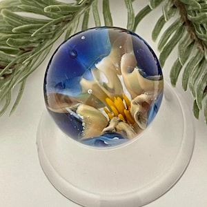 May include: A clear glass paperweight with a spherical shape. The interior features a design with blue, yellow, and tan colors, resembling a flower. The paperweight is set on a clear, circular base, with a green leafy branch in the background.