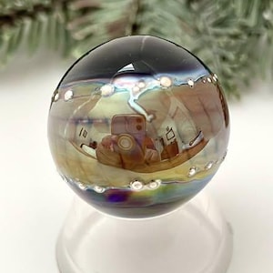 May include: A clear, iridescent glass orb with a dark top and a ring of small, white, pearl-like accents. The orb reflects the surrounding environment, sitting on a clear, rounded base. The background features blurred green foliage.