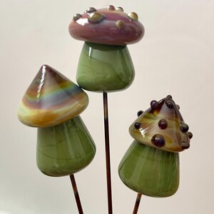 May include: Three glass mushroom garden stakes. The mushroom caps are various colors, including purple, brown, and iridescent. The stems are green, and the stakes are brown metal. The mushrooms are a decorative garden accent.