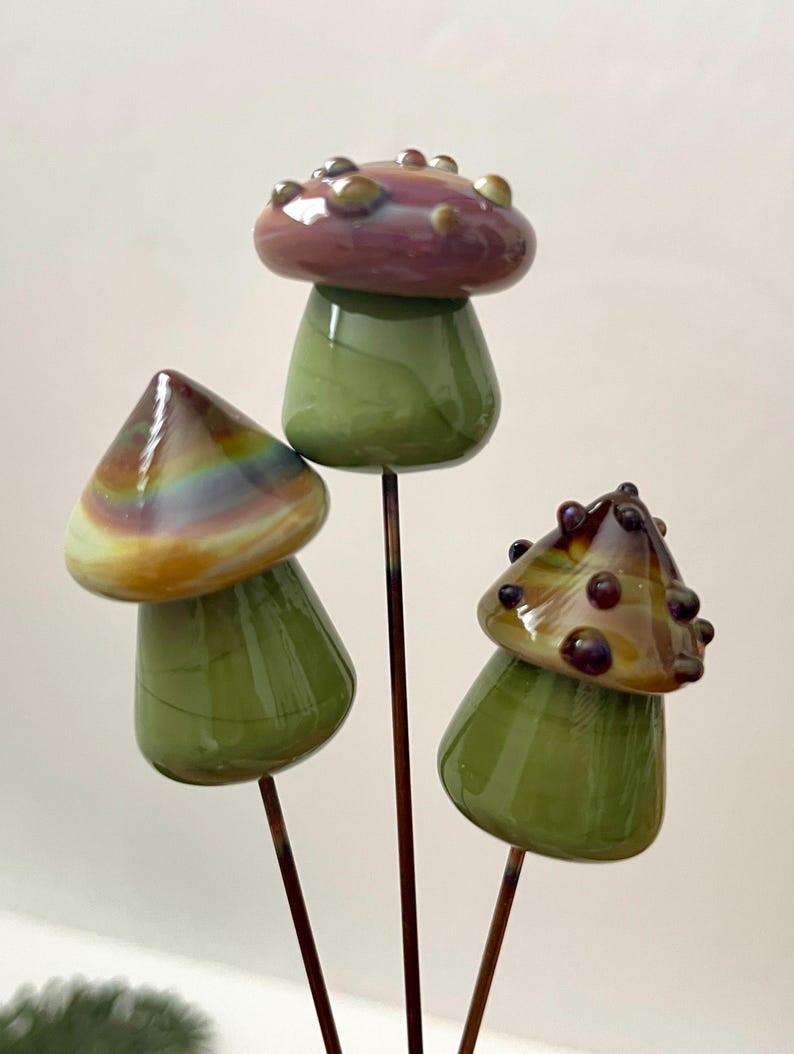 May include: Three glass mushroom garden stakes. The mushroom caps are various colors, including purple, brown, and iridescent. The stems are green, and the stakes are brown metal. The mushrooms are a decorative garden accent.