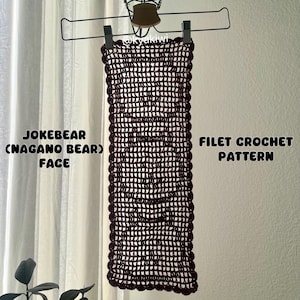 Könnte beinhalten: Ein dunkelburgunderfarbenes Filet-Häkelmuster wird gezeigt, das an einem Metallbügel hängt. Das Muster zeigt ein Bärengesicht-Design. Der Text links lautet "JOKEBEAR (NAGANO BEAR) FACE". Der Text rechts lautet "FILET CROCHET PATTERN".