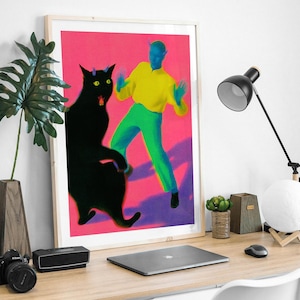 May include: A framed art print featuring a black cat and a figure in yellow and green against a pink background. The artwork is displayed on a wooden desk with a laptop, lamp, and decorative items. The print has a light wood frame.