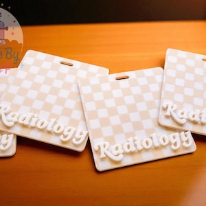 May include: Several cream-colored badge holders with a checkered pattern and the word "Radiology" in a 3D font. Each badge has a slot for a lanyard. The badges are arranged on a wooden surface.