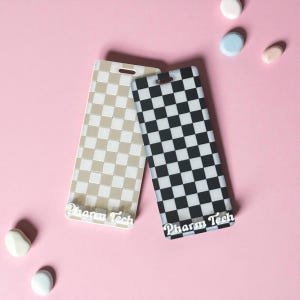 May include: Two rectangular phone cases with a checkered pattern. One case is beige and the other is black and white. The text "Pharm Tech" is printed on the bottom of each case. The cases are on a pink background.