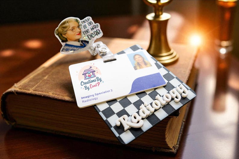 May include: A badge reel with a white and blue ID card, a photo ID, and a black and white checkered base with the word "Radiology". A small charm with the text "Help is on the way dear" is attached.