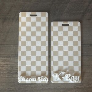 May include: Two rectangular, off-white and tan checkered badge holders. One reads "Pharm Tech" in white script, the other reads "X-Ray". Each has a slot at the top for a lanyard.