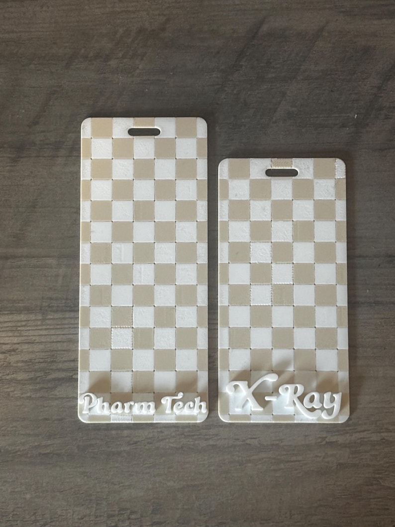 May include: Two rectangular, off-white and tan checkered badge holders. One reads "Pharm Tech" in white script, the other reads "X-Ray". Each has a slot at the top for a lanyard.