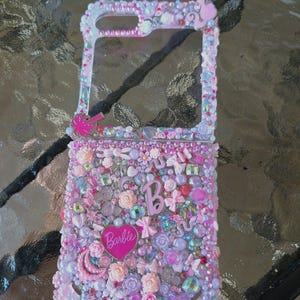 May include: A bedazzled phone case with a pink, white, and purple color scheme. The case is covered in various embellishments, including flowers, beads, and the Barbie logo. The case has a clear window for the phone's screen.