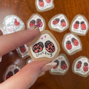 May include: A collection of small, irregular-shaped stickers. Each sticker features two red ladybugs with black spots and the words "love bug" in black script. The stickers have a clear, glossy finish and are on a brown surface.
