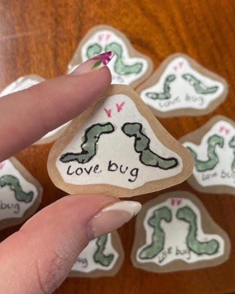 May include: A collection of small, light-colored, irregularly shaped cookies, each decorated with a green worm-like design and the words "Love bug" in black. One cookie is held by a person's hand.