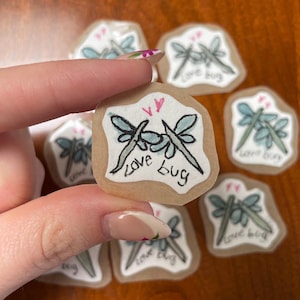 May include: A collection of small, handmade stickers featuring a dragonfly design. Each sticker has a light brown background with a pair of dragonflies in black and light blue, with the words "love bug" written below. Two pink hearts are above the dragonflies.