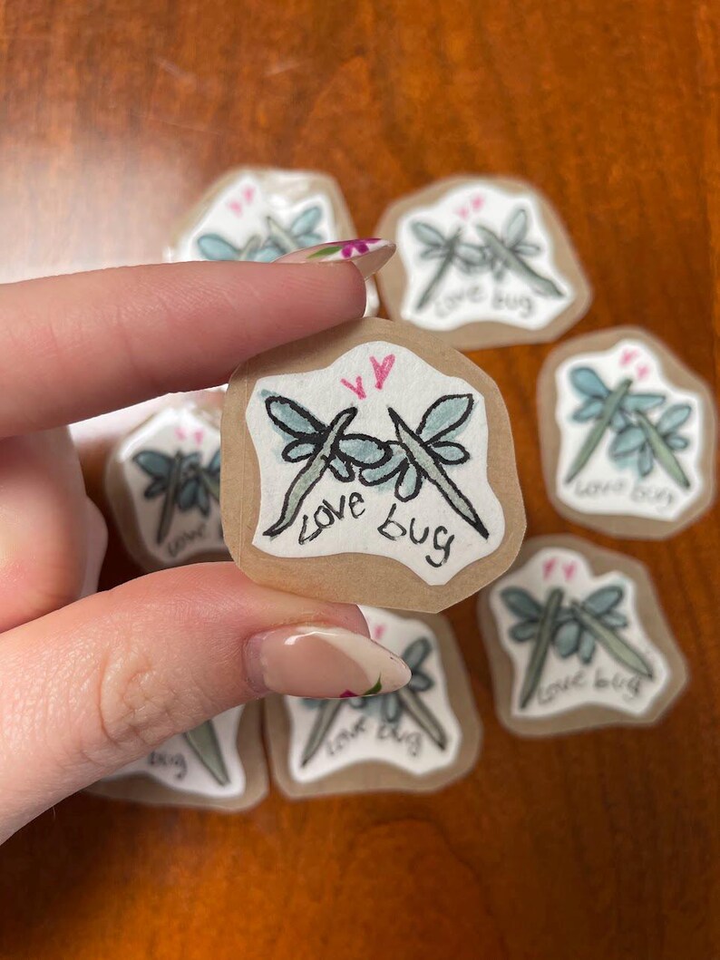 May include: A collection of small, handmade stickers featuring a dragonfly design. Each sticker has a light brown background with a pair of dragonflies in black and light blue, with the words "love bug" written below. Two pink hearts are above the dragonflies.