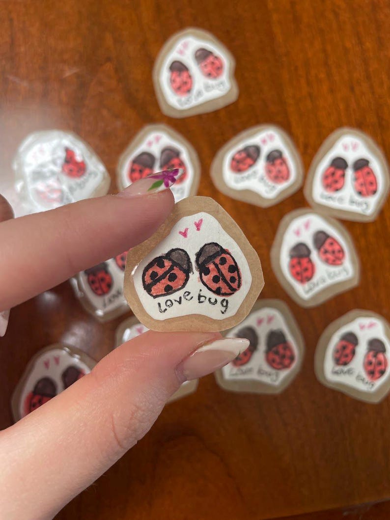 May include: A collection of small, irregular-shaped stickers. Each sticker features two red ladybugs with black spots and the words "love bug" in black script. The stickers have a clear, glossy finish and are on a brown surface.