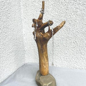 Puede incluir: Expositor de joyas artesanal hecho de madera flotante. Montado sobre una base de piedra gris, exhibe varios anillos y pendientes colgantes plateados. La madera tiene un color marrón cálido natural.