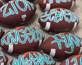 May include: Several brown American footballs with white stripes, each bearing a name in teal script. Names visible include Connor, Zion, Harley, Sebastian, Kingston, and Nathaniel. The footballs have an orange stripe.
