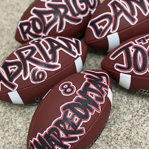 May include: A collection of personalized brown footballs with white stripes. Each football features a name in black and pink graffiti-style lettering. One football displays the number 8.