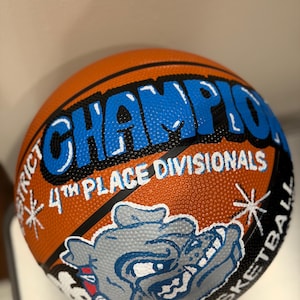 May include: An orange basketball with black seams and blue and white text that reads "CHAMPION" and "4th PLACE DIVISIONALS". A gray bulldog graphic is also featured on the ball.