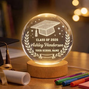 May include: A glowing graduation-themed crystal ball lamp with the text "Class of 2026 Ashley Henderson Your School Name". The lamp sits on a wooden base and features a graduation cap design.