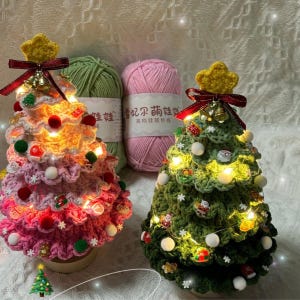 May include: Two handmade crochet Christmas trees, one pink and one green, are illuminated with fairy lights. Each tree is adorned with small ornaments and topped with a gold star and a red bow. Behind the trees are two skeins of yarn, one green and one pink.