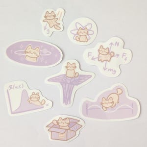 May include: A collection of eight stickers featuring cartoon cats in various poses and settings. The stickers are primarily light purple and beige, with designs including a cat in space, on a graph, and in a box. The stickers are die-cut.