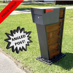 May include: A gray metal mailbox with a red flag and a locking door, mounted on an angled post featuring horizontal dark wood paneling. The number 5400 is displayed vertically in black digits on the wood. The base is surrounded by black stones.