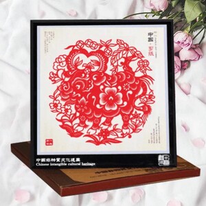 May include: A framed red paper cut-out artwork featuring a detailed floral and animal design. The artwork is set against a white background and framed in black. The text "Chinese intangible cultural heritage" is visible on the frame.
