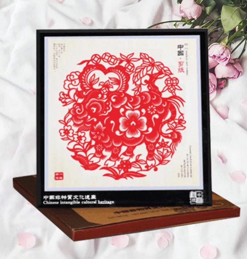 May include: A framed red paper cut-out artwork featuring a detailed floral and animal design. The artwork is set against a white background and framed in black. The text "Chinese intangible cultural heritage" is visible on the frame.