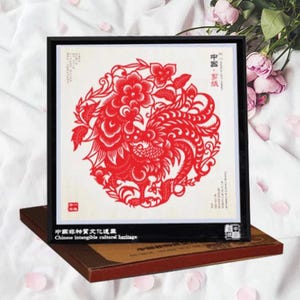 May include: A framed red paper cut art piece featuring a phoenix design. The artwork is set against a white background and framed in black. The text "Chinese intangible cultural heritage" is visible.