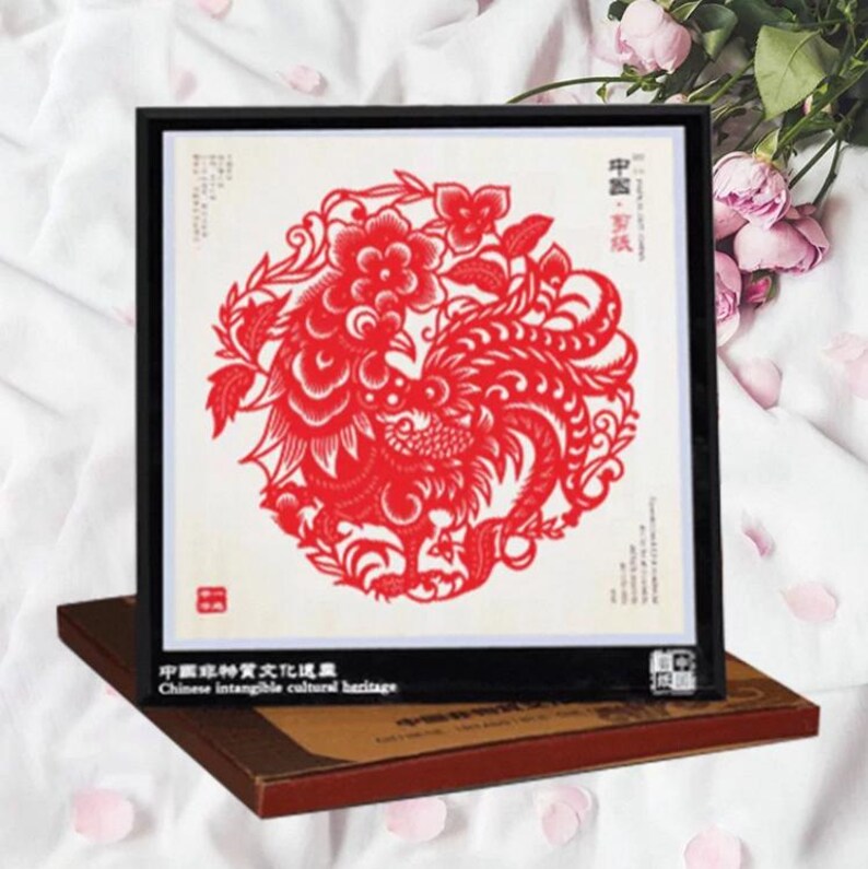 May include: A framed red paper cut art piece featuring a phoenix design. The artwork is set against a white background and framed in black. The text "Chinese intangible cultural heritage" is visible.