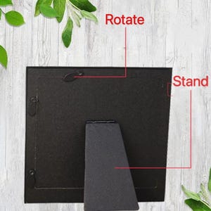May include: A black picture frame with a stand and a rotating mechanism. The word "Rotate" is written above the frame, and the word "Stand" is written to the right. The frame is set against a light wood-grain background.