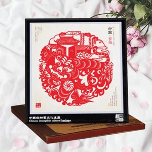 May include: A framed red paper cut art piece with a detailed circular design featuring buildings and swirling patterns. The frame is black, and the artwork rests on a brown box. Text in Chinese and English is visible.