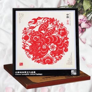 May include: A framed red paper cut-out artwork featuring a detailed floral and animal design. The artwork is set against a white background and framed in black. The text "Chinese intangible cultural heritage" is visible.