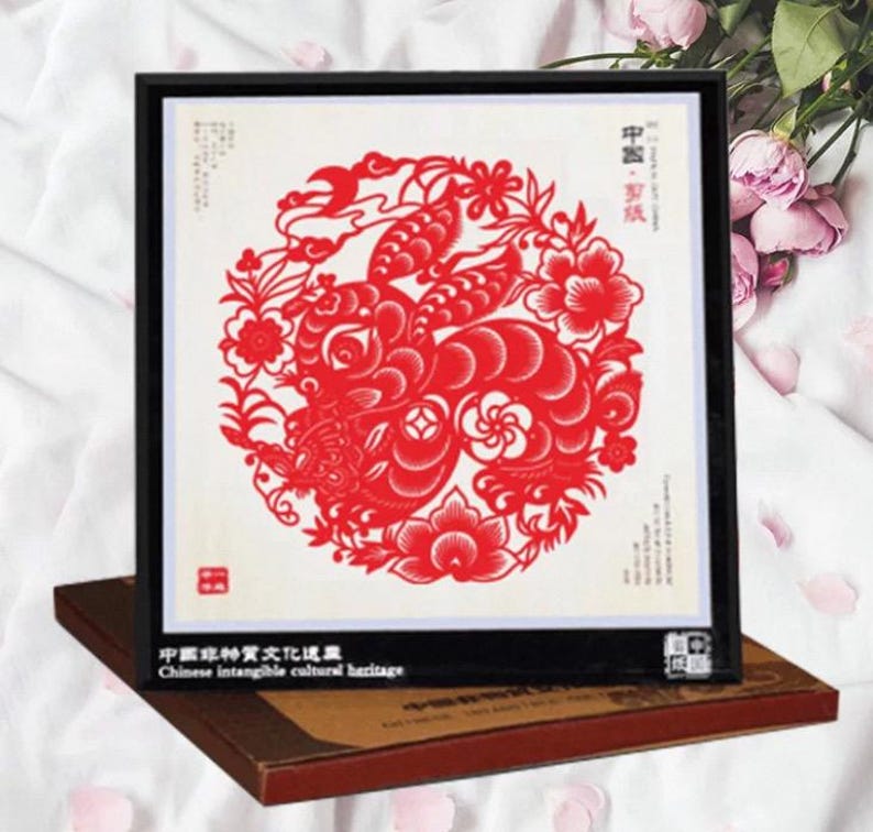May include: A framed red paper cut-out artwork featuring a detailed floral and animal design. The artwork is set against a white background and framed in black. The text "Chinese intangible cultural heritage" is visible.